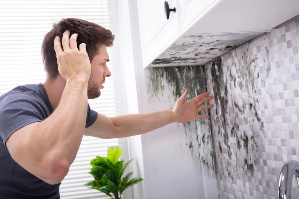 Homme touchant le mur au-dessus d'un évier, montrant des traces noires qui descendent du plafond, illustrant les dégâts constatés dans le logement