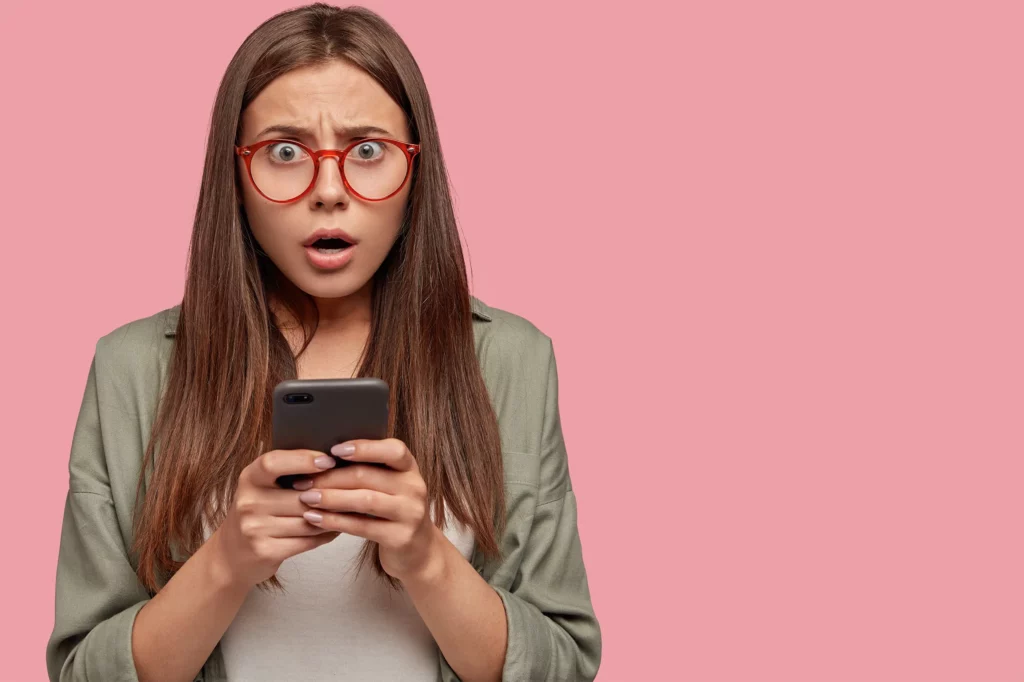 Femme avec des lunettes tenant un téléphone portable entre les deux mains, regardant la caméra avec une expression de surprise et la bouche ouverte, illustrant la réaction à la réception ou la lecture d'un SMS