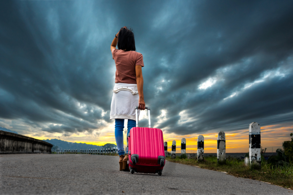 Femme tenant une valise à roulette, marchant sur une route de campagne bordée de plots, sous un ciel nuageux et menaçant, illustrant l'abandon du domicile conjugal