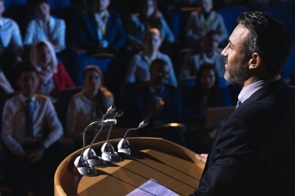 Homme en costume vu de dos, debout à un pupitre avec des micros, s'adressant à une assemblée de personnes