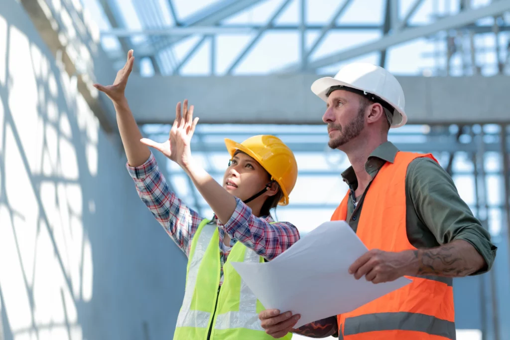 Couple de professionnels dans un immeuble en construction, tous deux portant des casques de chantier. La femme avance ses mains vers un mur pour visualiser quelque chose, tandis que l'homme tient un papier et observe les mains de la femme pour vérifier les détails.