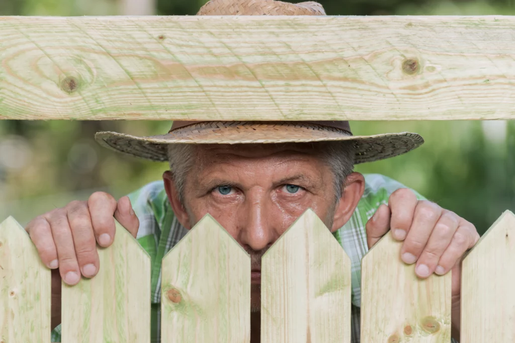 Homme portant un chapeau de paille, regardant par-dessus une clôture pour espionner le voisin, illustrant un conflit de voisinage et la problématique de l'intrusion ou de l'envahissement de la vie privée