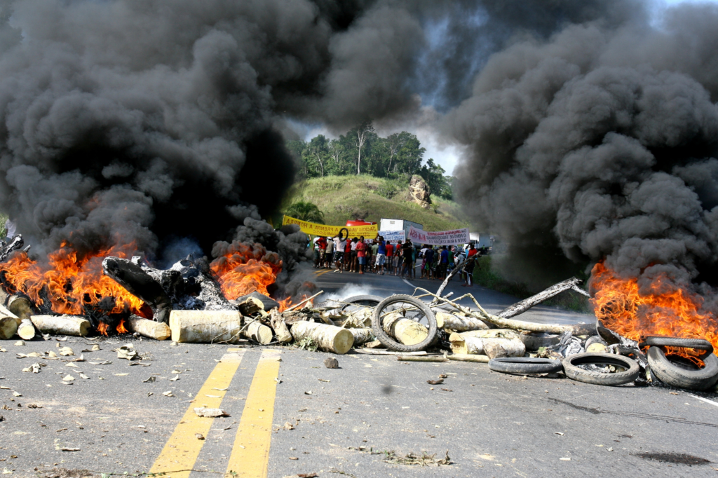 Chaussée goudronnée avec une barricade en feu composée de pneus et de différents matériaux, et des manifestants en grève en arrière-plan