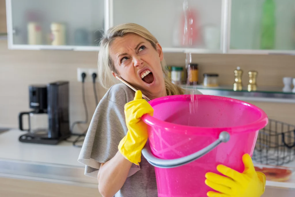 Femme portant des gants jaunes, tenant un sceau rose sous une fuite d'eau du plafond, tout en parlant au téléphone collé à son oreille, illustrant la gestion d'un dégât des eaux et la nécessité de contacter un professionnel pour un constat d'huissier