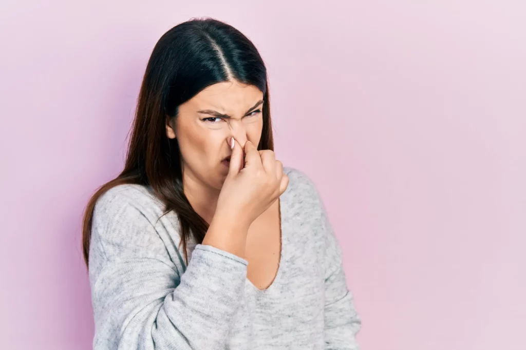 Femme se bouchant le nez avec une expression de gêne, sur fond rose, illustrant la gêne causée par une nuisance olfactive et la nécessité de documenter les problèmes d'odeurs pour un constat