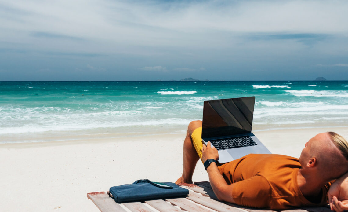 Homme allongé sur son lieu de villégiature, à savoir une plage, avec un ordinateur sur les génoux