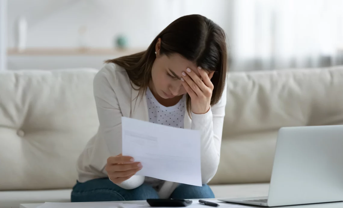 Femme assise sur son canapé tenant un papier pour illustrer la réception d'un courrier envoyé par la banque lui indiquant que son chèque a été rejeté.